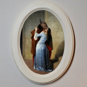 May include: A painting of a couple kissing in a doorway. The man is wearing a brown coat and a hat, and the woman is wearing a blue dress. The painting is framed in a white oval frame.