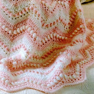 Può includere: Una coperta all'uncinetto fatta a mano, morbida e accogliente, nei toni del bianco e del rosa chiaro. La coperta presenta un motivo a zig-zag strutturato con un bordo smerlato. Ideale per un bambino o come coperta decorativa.