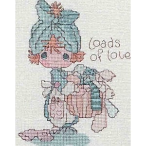 May include: A cross-stitch embroidery pattern featuring a character with orange hair wearing a large blue hat, carrying a bundle of gifts, with the text Loads of love stitched in brown thread on a light fabric background with visible weave texture.