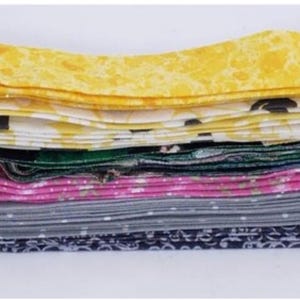Pode incluir: Uma pilha de tiras de tecido dobradas com vários padrões e cores. A tira superior é amarela com uma textura manchada, seguida por uma tira branca com formas pretas, uma tira verde-escura, uma tira rosa floral, uma tira cinza com bolinhas e uma tira preta floral.