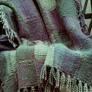 Op de afbeelding: Een zacht, geweven deken in groen en paars met franjes aan de randen.