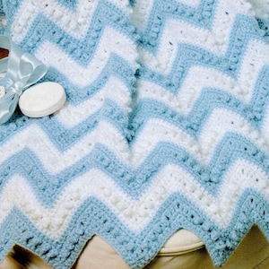 Könnte beinhalten: Eine handgefertigte, gehäkelte Decke mit Zickzackmuster in Weiß und Hellblau. Ein hellblaues Band und ein weißes, rundes Objekt liegen auf der Decke. Die Ränder der Decke haben ein gewelltes Design.