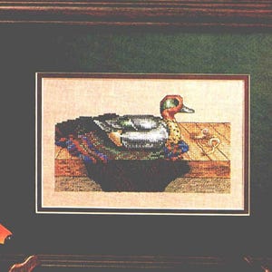 Puede incluir: Obra de arte en punto de cruz enmarcada de un pato. El pato tiene una cabeza verde, un pico naranja y un plumaje colorido. La obra de arte está enmarcada en un marco de madera marrón oscuro con un fondo verde.