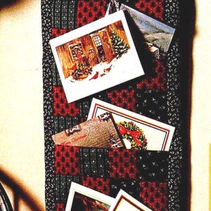 May include: A vertical fabric wall organizer featuring a black and red patterned quilt design with multiple pockets. The organizer includes a red and white polka dot ribbon hanger and holds several rectangular greeting cards with holiday imagery.