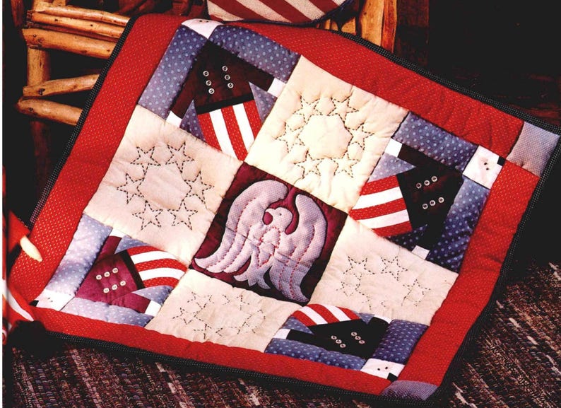 May include: A patriotic quilt with a red border and a patchwork design. The quilt features squares with stars, stripes, and an eagle motif in red, white, and blue. The quilt is likely handmade and evokes a sense of Americana.