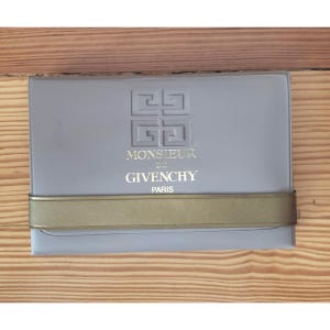 May include: A rectangular, pale grey case with a gold band. The case has an embossed logo and gold text reading "MONSIEUR DE GIVENCHY PARIS". It is likely designed to hold a fragrance or grooming product.