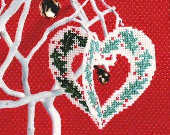 May include: A white, cross-stitched heart-shaped Christmas ornament with green and red accents, hanging from a white branch. The ornament has a hollow center and is set against a red dotted background. Small gold bells are also visible.