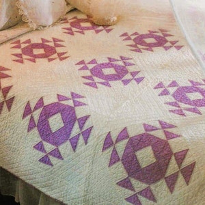 May include: A white quilt with a purple geometric pattern. The quilt is made up of repeating squares with a central diamond shape surrounded by triangles.