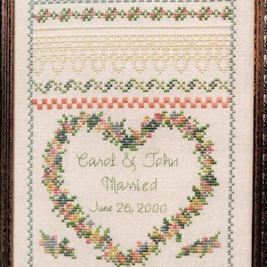 May include: A white cross-stitch sampler with a floral heart design. The heart is surrounded by a green and blue floral border. The text inside the heart reads "Carol & John Married June 26, 2000".