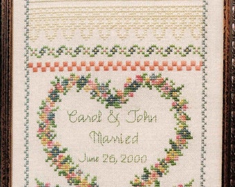 Puede incluir: Un sampler de punto de cruz blanco con un diseño de corazón floral. El corazón está rodeado por un borde floral verde y azul. El texto dentro del corazón dice "Carol & John Married June 26, 2000".