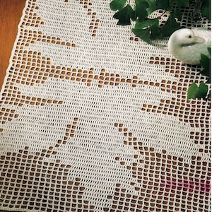 May include: Cream-colored, open-weave crochet table runner with a floral design. The runner is rectangular and features a repeating pattern of stylized flowers and leaves. The crochet work is detailed and creates a delicate, textured appearance.