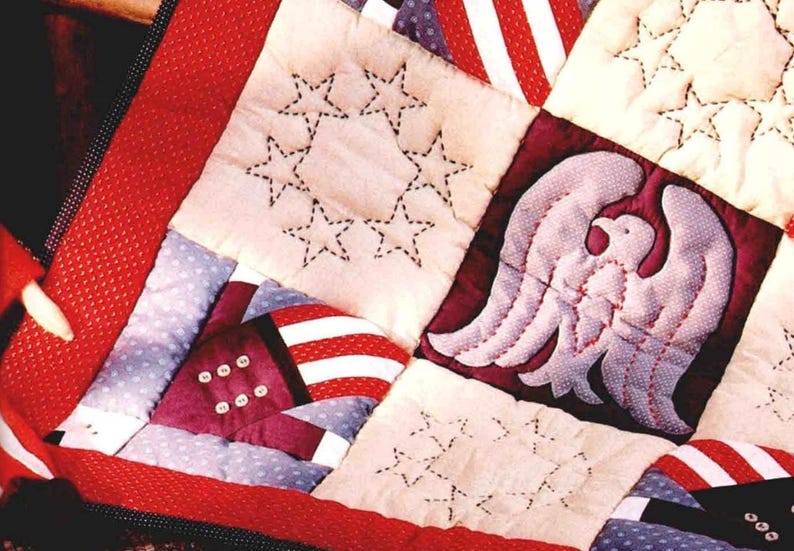 May include: Close-up of a handmade quilt with a patriotic theme. The quilt features squares with embroidered stars, stripes, and an eagle design. The color palette includes red, white, and blue, with a stitched border.