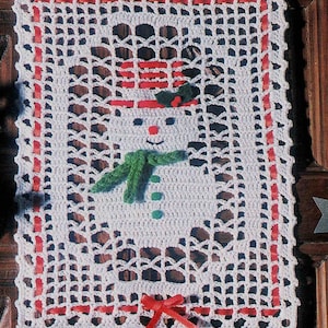 May include: White crocheted doily with a red border and a snowman design in the center. The snowman has a red and white striped hat, a green scarf, and a green hat.