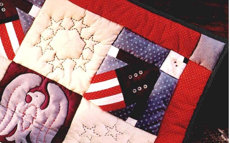 May include: Close-up of a quilted patchwork with patriotic themes. The quilt features red and white stripes, blue polka dots, and a white star pattern. An embroidered eagle is also visible. The quilt is bordered with a red fabric.
