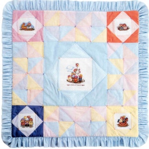 May include: A square patchwork quilt featuring a light blue ruffled border and a grid of pastel gingham triangles. Five white fabric squares are appliqued onto the quilt, each displaying a cross-stitched scene of dolls. The centre square includes the text LOVE LIFTED ME.