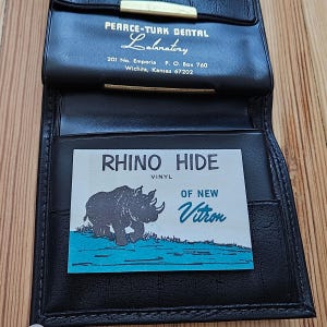 May include: Black leather wallet with a gold clasp, open to reveal a card that reads "RHINO HIDE VINYL" with a rhino illustration. The wallet also has text that reads "PEARCE-TURK DENTAL Laboratory".