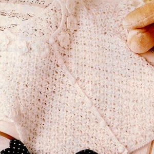 May include: A white crocheted cardigan featuring a delicate openwork diamond pattern, a round neckline, and a front button closure with small matching white buttons. The garment is adorned with a row of small, textured crocheted flower embellishments.