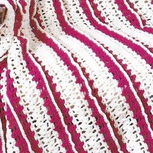 Könnte beinhalten: Nahaufnahme einer gehäkelten Decke mit abwechselnden weißen und magentafarbenen Streifen. Das Häkelmuster weist ein strukturiertes Design mit offenen Räumen auf, wodurch eine optisch ansprechende und gemütliche Textur entsteht. Die diagonale Ausrichtung der Decke fügt ein dynamisches Element hinzu.