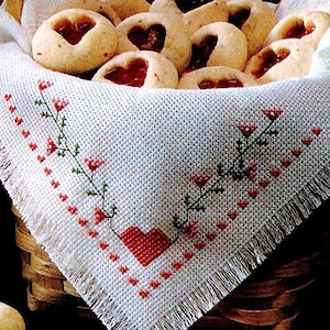 May include: A white crocheted doily with a red heart design and a border of red flowers. The doily is in a wicker basket filled with heart-shaped cookies.