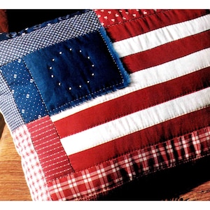May include: A patriotic pillow with a patchwork design resembling the American flag. The pillow features red and white stripes, a blue square with white stars, and various patterned fabric borders. The overall aesthetic is a vintage, handcrafted style.