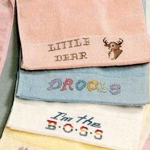 May include: A stack of embroidered towels in pastel shades. The top towel is pink with "LITTLE DEAR" and a deer. Below are blue, white, and yellow towels with embroidered text: "DROOLS" and "I'm the B.O.S.S."