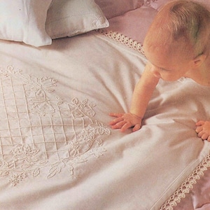 Puede incluir: Una manta de bebé blanca con un borde de ganchillo y un diseño de corazón bordado. Un bebé está gateando sobre la manta.