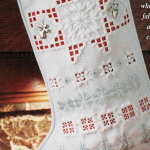 May include: A white Christmas stocking featuring the word REJOICE embroidered in silver thread. The fabric displays intricate cutwork, red square accents, silver stitched pine trees, and two small silver angel charms attached to the front.