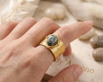 May include: A wide gold-toned ring featuring a textured, ribbed band and a central oval-shaped stone with a mottled grey and black pattern. The stone is held in place by a decorative metal setting with small bead accents on the top and bottom.