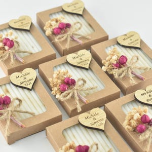May include: Kraft paper boxes with a clear window, each containing a white item and a small bouquet of dried flowers and pink buds. A wooden heart with the names "Michèle & Simone 7.9.24" is attached to each box, suitable for wedding favors.