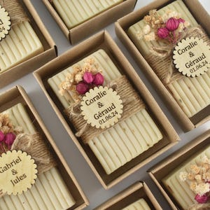 May include: A collection of small, rectangular boxes wrapped in brown paper and twine. Each box contains a bar of soap with dried flowers and a personalized wooden tag. The tags include the names "Coralie & Géraud" and "Gabriel & Jules" with a date.