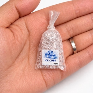 May include: A miniature bag of clear ice cubes, held in a hand. The bag is transparent, allowing the ice cubes to be visible. A label on the bag reads "ICE CUBE" with a graphic of blue ice cubes.
