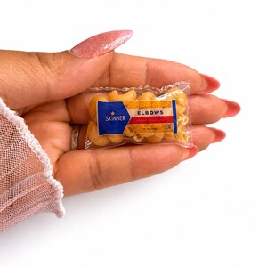 May include: A miniature clear plastic bag filled with small, dry, tan-colored elbow macaroni pasta. The front of the bag features a blue, white, and red label with the text Skinner, a star icon, and the word ELBOWS printed in bold black letters.