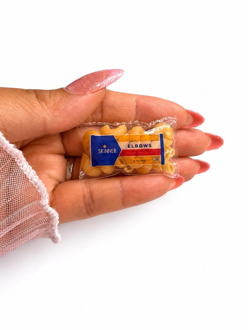 May include: A miniature clear plastic bag filled with small, dry, tan-colored elbow macaroni pasta. The front of the bag features a blue, white, and red label with the text Skinner, a star icon, and the word ELBOWS printed in bold black letters.