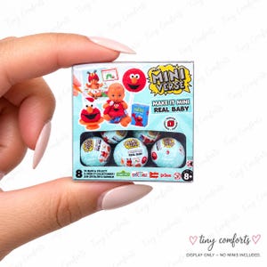 May include: A square MGA's Miniverse Make It Mini Real Baby Series 1 box featuring Elmo graphics, a baby doll illustration, and five light blue mystery spheres visible through a cutout window. The packaging includes text branding and age 8+ label.