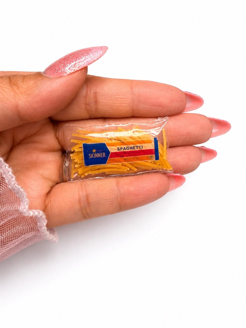 May include: A miniature clear plastic packet filled with small yellow pasta strands. The label on the front features a blue rectangle with a gold star and the text SKINNER, followed by a red and orange striped section with the word SPAGHETTI.