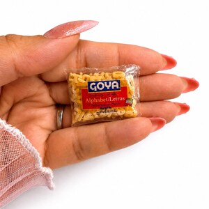 May include: A miniature clear plastic packet filled with small, beige, letter-shaped pasta. The label features the blue Goya logo above a red rectangular field with the white text Alphabet/Letras, along with additional small printed details.