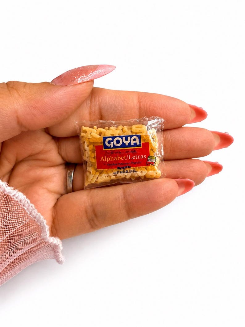May include: A miniature clear plastic packet filled with small, beige, letter-shaped pasta. The label features the blue Goya logo above a red rectangular field with the white text Alphabet/Letras, along with additional small printed details.