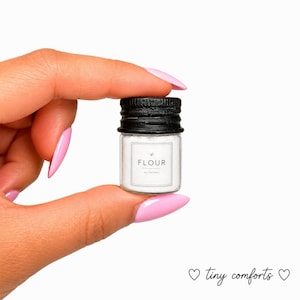May include: A miniature clear glass jar filled with white powder, featuring a black screw-top lid and a white label with a thin black border. The label displays the text FLOUR and ALL NATURAL in a clean, sans-serif font with a small leaf icon.