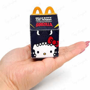 May include: A miniature black cardboard box with a yellow handle, featuring the text Hello Kitty and Friends Godzilla. The front displays a graphic of Hello Kitty wearing a black Godzilla costume with a red bow, set against a dark background.