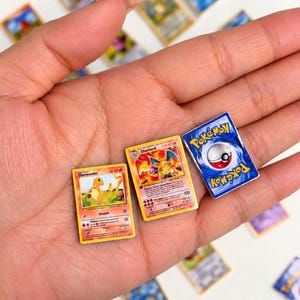 Puede incluir: Tres réplicas en miniatura de cartas coleccionables que incluyen una carta de personaje Charmander, una carta de Charizard y una carta Pokémon con el reverso azul. Las cartas son rectangulares con ilustraciones coloridas.