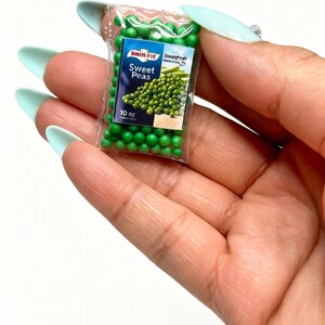 K&ouml;nnte beinhalten: Miniaturbeutel mit Birds Eye Sweet Peas, ca. 283 Gramm. Der transparente Plastikbeutel enth&auml;lt kleine gr&uuml;ne Erbsen und ein Etikett mit dem Birds Eye-Logo und den Worten "Sweet Peas". Der Beutel wird in einer Hand mit hellblauen N&auml;geln gehalten.