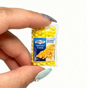 K&ouml;nnte beinhalten: Miniaturbeutel mit Birds Eye Super Sweet Corn. Der transparente Plastikbeutel enth&auml;lt gelbe Maisk&ouml;rner und hat ein blau-wei&szlig;es Etikett. Auf dem Etikett steht "Birds Eye" und "Super Sweet Corn". Der Beutel wird zwischen Fingern gehalten.