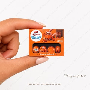 May include: A rectangular orange Zuru My Mini Baby Pumpkin Patch toy box featuring a graphic of a baby inside a pumpkin. The packaging includes the text My Mini Baby and Pumpkin Patch, with a clear window displaying four small orange spheres.