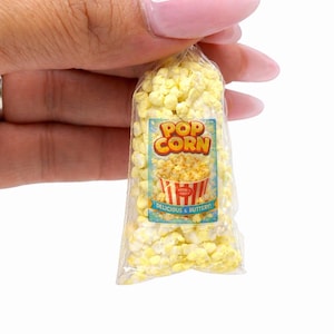 May include: A miniature clear plastic bag filled with small yellow popcorn kernels. The front of the bag features a label with the text POP CORN in orange letters, an illustration of a striped bucket, and the text DELICIOUS & BUTTERY.