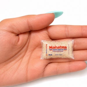 May include: A miniature clear plastic bag filled with white rice grains, featuring the red and blue Mahatma logo, the text America's Favorite Rice, Extra Long Enriched, and a net weight of 1 oz (28.3 g) printed on the front of the small package.
