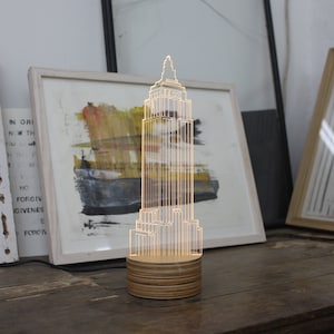 May include: A clear acrylic skyscraper lamp with a wooden base. The lamp is lit up and casting a warm glow. The lamp is sitting on a wooden table.