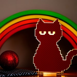 Op de afbeelding: Een rood gepixelde kattennachtlampje met een witte omtrek, voor een regenboogkleurige houten boog.