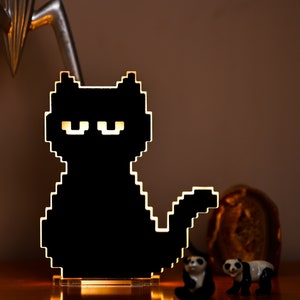 May include: A black pixelated cat night light with a white base. The cat is sitting with its tail curled around its body. The light is on and illuminating the cat's eyes.