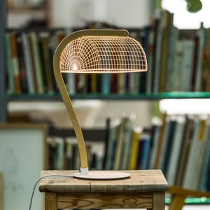 May include: A wooden desk lamp with a geometric wire shade. The lamp is on a wooden table and is lit, casting a warm glow. The lamp is in a room with bookshelves and a chair.
