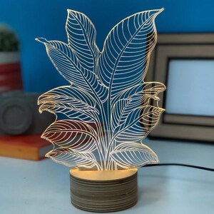 May include: A white acrylic night light shaped like a leafy plant with a wooden base. The light is illuminated and casts a warm glow.